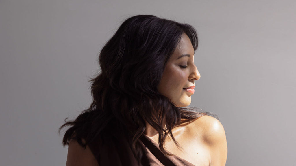 Woman sitting on a white surface wearing a brown top and blue jeans against a plain background